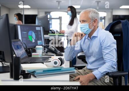 Nachdenklicher Senior Geschäftsmann mit Gesichtsmaske auf seinem Schreibtisch sitzend Im modernen Büro Stockfoto