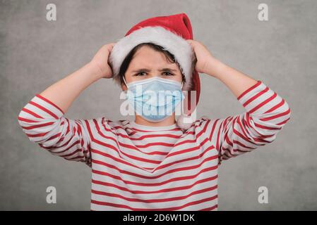 Frohe Weihnachten, wütend Kind mit medizinischen Maske trägt Weihnachtsmann Hut Mit den Händen auf dem Kopf Stockfoto