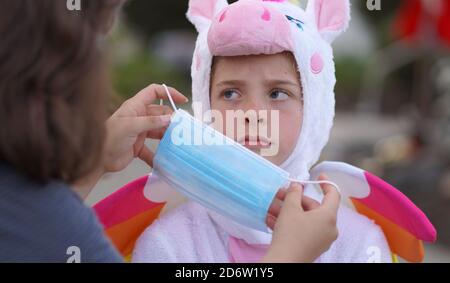 Ein junges Mädchen in einem Halloween Kostüm sieht sehr unglücklich aus, als ihre Mutter eine Gesichtsmaske auf sie legt, um ihre Sicherheit und Schutz in Zeiten von Covid-19. Stockfoto