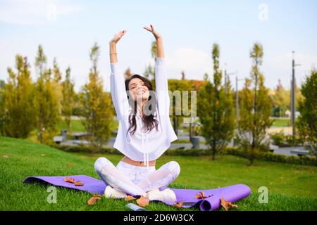 Attraktive Brünette Mädchen in weißen Sportbekleidung sitzen auf lila Matte in Lotus-Pose und strecken sich die Hände unter dem Kopf, Outdoor-Fitness, Yoga-Übungen Stockfoto