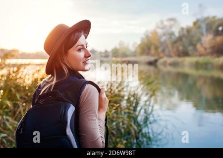 Reisende mit Rucksack entspannen am Herbstfluss bei Sonnenuntergang. Junge Frau genießt Herbstwetter und Landschaft Stockfoto