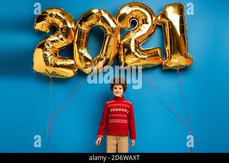 Fröhlicher Junge in roten Pullover stehen neben goldenen Luftballons mit 2021 Zahlen auf blau Stockfoto