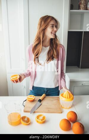 Schöne Ingwer Frau mit Sommersprossen lächelt beim Schneiden Orangen Für die Herstellung von Saft mit einer manuellen Quetschpresse Stockfoto