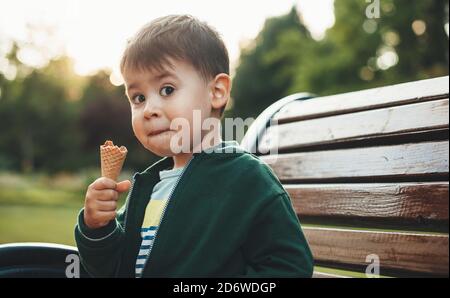 Netter Junge isst Eis und schaut überrascht an Kamera, während Sie auf der Bank im Park sitzen Stockfoto