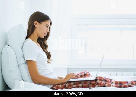 Seitenansicht einer jungen Frau, die im Schlafzimmer auf dem Bett sitzt und auf dem Laptop tippt. Arbeiten Sie von zu Hause aus Stockfoto