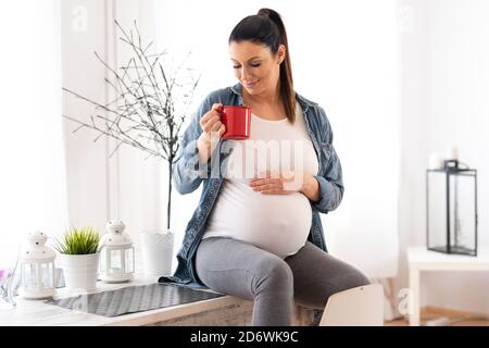 Ein schönes Lächeln schwangere Frau sitzt auf einem Küchentisch in einer Jeansjacke und trinken einen Kaffee aus einer roten Tasse. Stockfoto