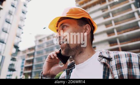 Porträt eines jungen Handwerkers, der beim Stehen auf der Baustelle anruft. Techniker, der auf einer Baustelle telefoniert. Builder in Helm Stockfoto