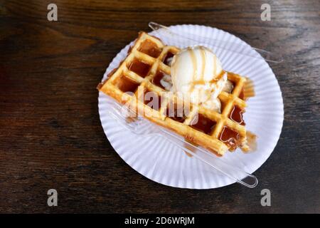 Dessert Belgische Waffel mit einem Ball von cremigem Eis gegossen mit Karamellsirup auf einem hölzernen Hintergrund, in einem Straßencafé auf dem Weihnachtsmarkt. Kontra Stockfoto
