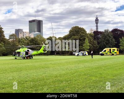 Ein krankes Kind wird aus der Kinderluftambulanz entfernt und in einen Krankenwagen im Regent's Park im Zentrum von London gebracht. Der Patient wird zum nahe gelegenen Great Ormond Street Hospital gebracht. Der Hubschrauber wird von Wohltätigkeitsorganisationen finanziert und wird verwendet, um Kinder zu bewegen, die zu krank sind, um auf der Straße zu reisen. Ein Polizist räumte das Feld von Menschen, bevor der Hubschrauber landete. Die drei Transportformen sind Millionen Pfund wert und der Service ist für den Patienten nach den NHS-Regeln kostenlos. Der BT Tower ist im Hintergrund. Stockfoto