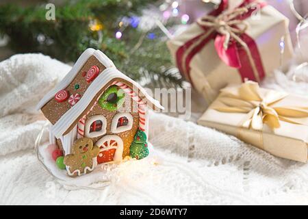 Schachteln mit Weihnachtsgeschenken und Dekorationen Lebkuchenhaus auf einer weißen Strickdecke in den Lichtern von Girlanden und Fichtenzweigen. Neujahr, festi Stockfoto