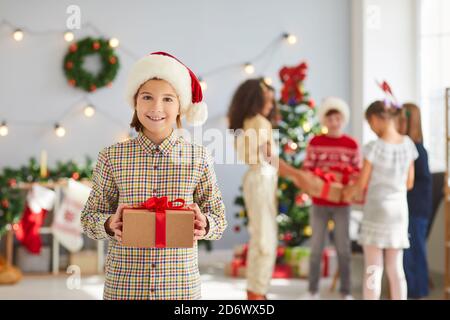 Glücklicher kleiner Junge in einem roten Weihnachtsmann Hut hält eine Geschenkbox in Kraftpapier und rotem Band verpackt. Stockfoto