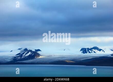 Schöne arktische Aussicht - schmelzender Gletscher, schwarze Berge mit Schnee bedeckt und blauer Golf vor dem Hintergrund der dramatischen bewölkten Himmel in der Nähe von Barentsburg, S Stockfoto