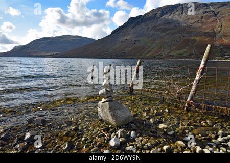 Ein Felsenkairn steht an der Küste von Wastwater im Lake District. Berge rund um den See und ein Metallzaun mit Holzpfosten erstrecken sich im See. Stockfoto