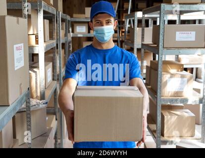 Männlicher Kurier trägt Gesichtsmaske hält Paketkasten im Lager, Porträt. Stockfoto