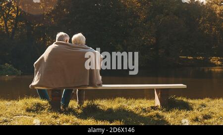 Ältere grauhaarige Paar sitzt auf der Bank in der Nähe des Flusses bedeckt mit Decke am Herbsttag. Romantik und Alter. Hochwertige Fotos Stockfoto