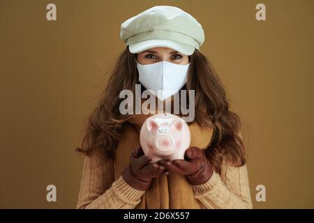 Leben während der covid-19 Pandemie. Portrait von eleganten mittleren Alters Hausfrau in Schal mit Sparschwein und weiße medizinische Maske isoliert auf beigem Hintergrund. Stockfoto