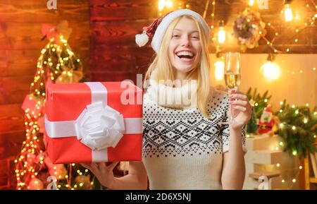 Glückliche blonde Frau in Santa roten Hut hält ein Geschenk Geschenk in einer Hand und eine Witwe Champagner in einer anderen In wunderschön dekorierten Innenraum Stockfoto