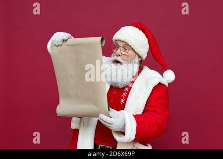 Schockiert Weihnachtsmann trägt Kostüm Lesung Wunschliste auf rotem Hintergrund. Stockfoto