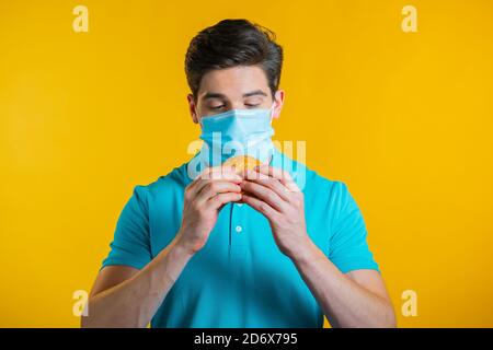 Junger Mann in schützende medizinische Maske essen Burger auf gelben Studio-Hintergrund. Guy macht Spaß, zeigt, wie man während einer Epidemie lebt. Stockfoto