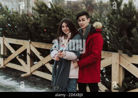 Ein nettes schönes Paar von einem Mann und einer Frau auf dem Hintergrund des Weihnachtsbaum Basar in der Stadt feiert das neue Jahr und klatscht einen pneumatischen Klapper mit Konfetti. Stockfoto