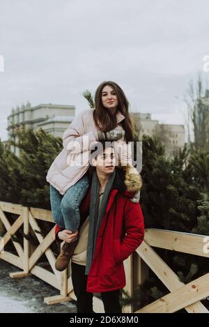 Eine junge Frau sitzt auf den Schultern eines Mannes vor dem Hintergrund von Weihnachtsbäumen und hat Spaß. Winterporträt von Liebenden. Stockfoto