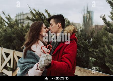 Eine junge Frau und ein Mann halten herzförmige Karamellstöcke und küssen vor dem Hintergrund des Weihnachtsbaum-Basars in der Stadt. Stockfoto