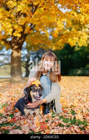 Glückliche junge kaukasische Frau im Herbst sitzen gelbe Blätter umarmen ihren Hund. Beste Freunde, die Spaß im Freien haben. Stockfoto