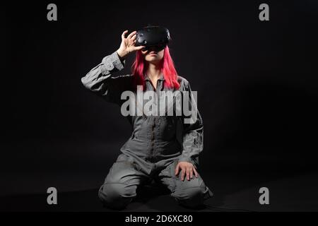 Junge Frau mit rosa Haaren und in einem Jumpsuit verwendet Virtual-Reality-Brille, ist im Spiel, schwarzer Hintergrund. VR-Simulation. Stockfoto