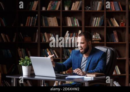 Wütend verrückt verärgerten Geschäftsmann in der Bibliothek Büro zeigt den Mittelfinger auf dem Laptop-Bildschirm. Anstrengende Verhandlungen durch Videokonferenz Stockfoto