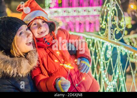 Kleiner Junge und seine Mutter mit Wunderkerzen in der Nähe von riesigen Tannenbaum und Weihnachtsbeleuchtung auf Weihnachtsmarkt. Weihnachten Feiertage auf fair Stockfoto