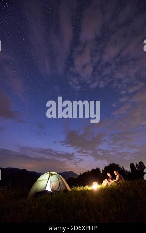 Silhouette von Touristen, die am Abend in den Bergen in der Nähe beleuchteten Zelt und ein Lagerfeuer unter schönem Himmel voller Sterne verbringen. Nachtcamping in den Bergen im Sommer Stockfoto