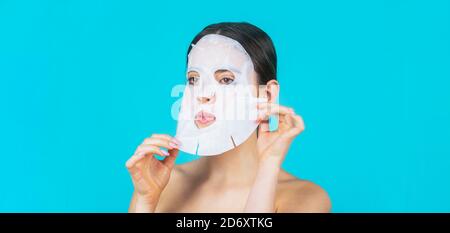 Feuchtigkeitsspendende Maske. Anti-Aging-Verfahren. Frau, die Blattmaske auf ihr Gesicht, auf blauem Hintergrund. Schöne Frau mit Maske Stockfoto