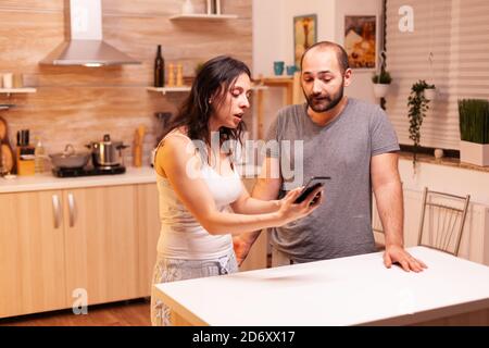 Frau schreit untreuen Mann in der Küche. Eifersüchtige Frau betrogen wütend frustriert beleidigt gereizt beschuldigen ihren Mann der Untreue zeigt ihm Nachrichten auf dem Smartphone schreien verzweifelt. Stockfoto