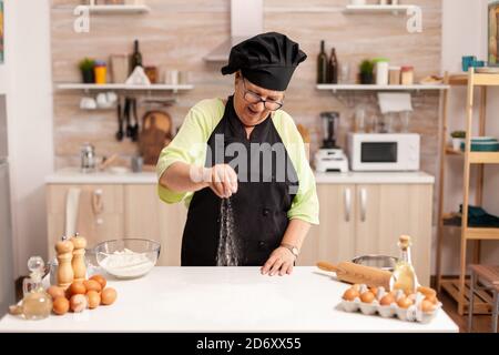 Die Chefköchin lächelte, während sie Pizza streute und Mehl auf den Küchentisch gab. Glücklicher älterer Koch mit einheitlicher Streuselung, sieben rohe Zutaten von Hand. Stockfoto