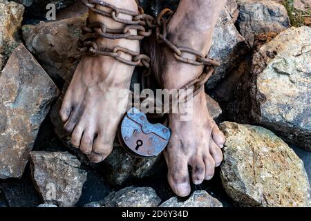 Die schmutzigen Füße des Sklaven sind in eine rostige Kette mit einem großen Schloss verwickelt. Sklavenarbeit Konzept Stockfoto