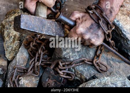 Die schmutzigen Hände eines Sklaven, der einen Hammer hält, sind in einer rostigen Kette mit einem großen Vorhängeschloss auf einem Hintergrund aus Steinen verwickelt. Stockfoto