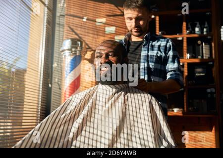 Männlicher Barbier, der beim Barbershop Umhang auf den Kunden legt Stockfoto