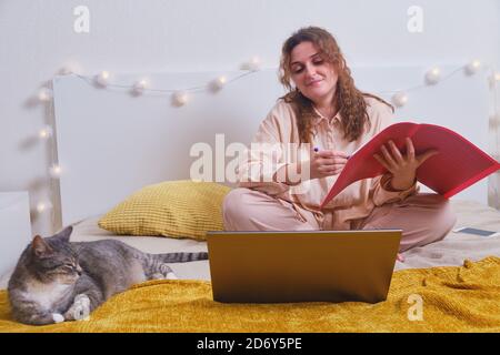 Glückliche Frau sitzt mit einem Laptop auf dem Bett und bekommt eine Ausbildung online. Katze und Mädchen während einer Lektion über das Internet, Lifestyle Stockfoto