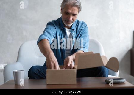 Lächelnder Mann Verbraucher offenen Karton erhalten Postpaket, männliche Kunde erhalten Kartonpaket sitzen auf Sofa zu Hause Stockfoto
