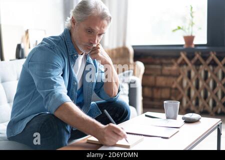 Mann in legerer Kleidung, der mit Finanzdokumenten arbeitet, während er zu Hause auf dem Sofa sitzt. Stockfoto