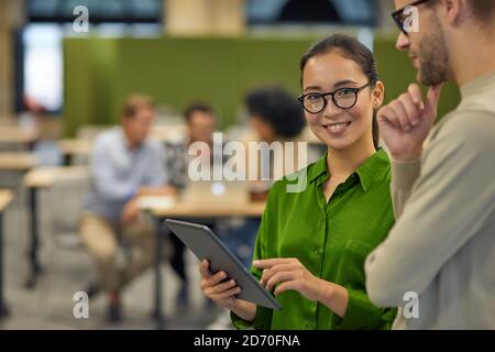 Zwei junge Coworker, Mann und Frau mit digitalem Tablet und diskutieren etwas, während sie im Coworking Space mit Kollegen im Hintergrund stehen. Geschäftsleute und moderne Technologien Stockfoto