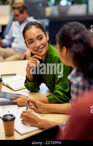 Vertikale Aufnahme einer jungen, fröhlichen Büroarbeiterin, die mit ihrer Kollegin etwas diskutiert, während sie am Schreibtisch sitzt und im Coworking Space oder Büro zusammenarbeitet. Business, Teamwork-Konzept Stockfoto