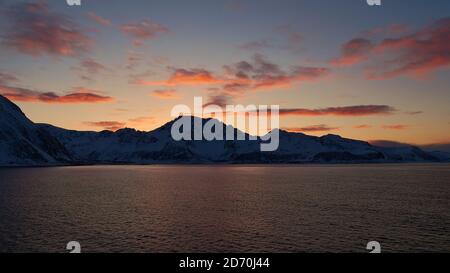 Atemberaubende Panoramaaussicht nach Sonnenuntergang auf schneebedeckte Berge an der norwegischen Küste, Skandinavien mit orangefarbenen Wolken, die sich im Wasser spiegeln. Stockfoto