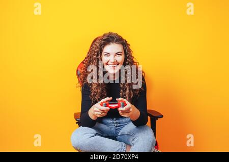 Frau, die hinter einem Spielstuhl in ihren Händen sitzt und rot hält Gamepad auf gelbem Hintergrund Stockfoto