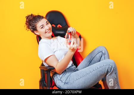 Frau, die hinter einem Spielstuhl in ihren Händen sitzt und rot hält Gamepad auf gelbem Hintergrund Stockfoto