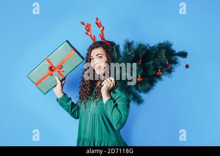 Junge Frau mit einem großen Geschenk und einem Weihnachtsbaum in den Händen, hat sie Hirschhörner auf ihrem Kopf auf einem blauen Hintergrund Stockfoto
