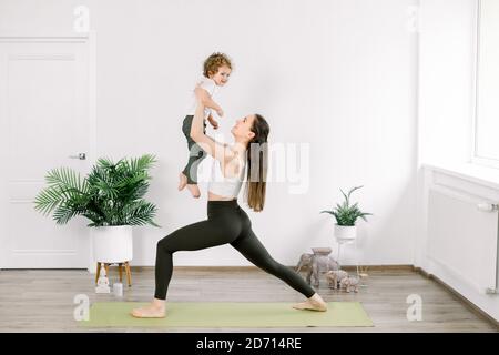 Mutter und kleiner Sohn in Sportbekleidung machen gemeinsam Yoga zu Hause. Schöne junge kaukasische Mutter praktiziert Yoga mit ihrem Sohn, Mutter in Yoga-Pose hält Stockfoto