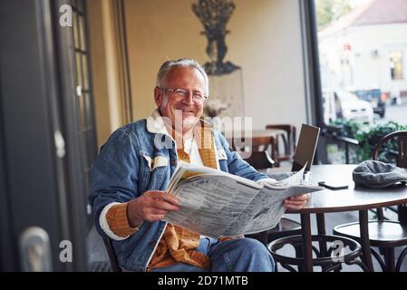 Stilvoller Senior in modischer Kleidung und Brille sitzt in Das Café und liest Zeitung Stockfoto