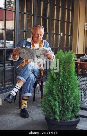 Stilvoller Senior in modischer Kleidung und Brille sitzt in Das Café und liest Zeitung Stockfoto