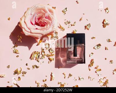 Rechteckige Parfümflasche aus Glas mit schwarzer Kappe, rosa Goldrose und Goldpapier auf einem pastellrosa Tisch. Luxuriöses Flatlay mit modischem Schatten. Stockfoto